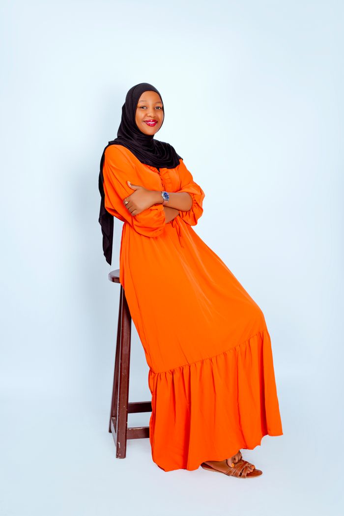 Stylish woman in a bright orange dress and hijab sitting confidently on stool against plain backdrop.