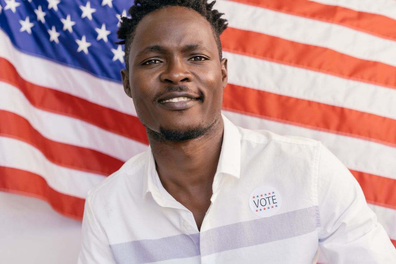 Adult man smiling with an 'I Voted' sticker and American flag backdrop.